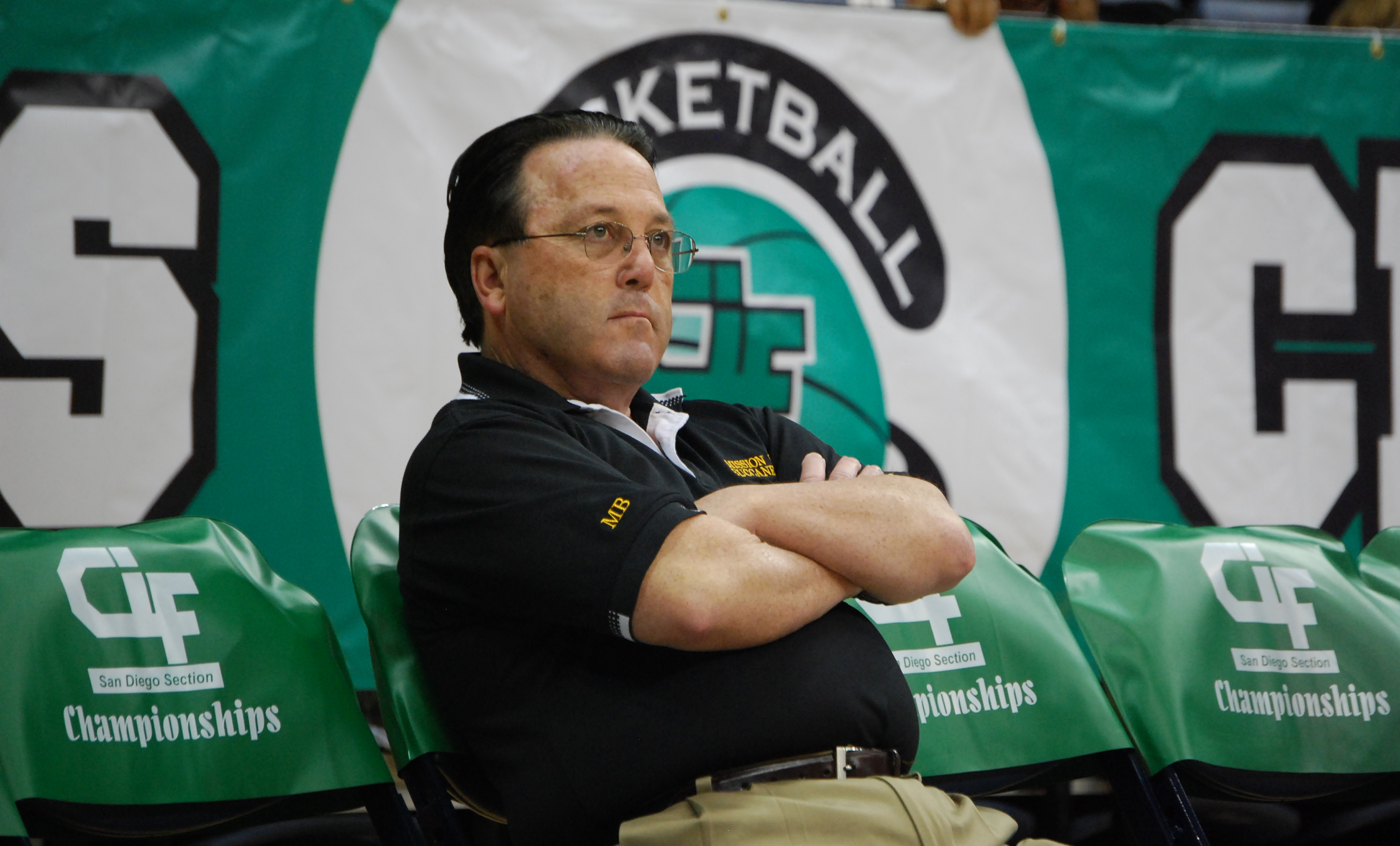 Coach Dennis Kane seated courtside with arms crossed in front of CIF San Diego Section Championship banners